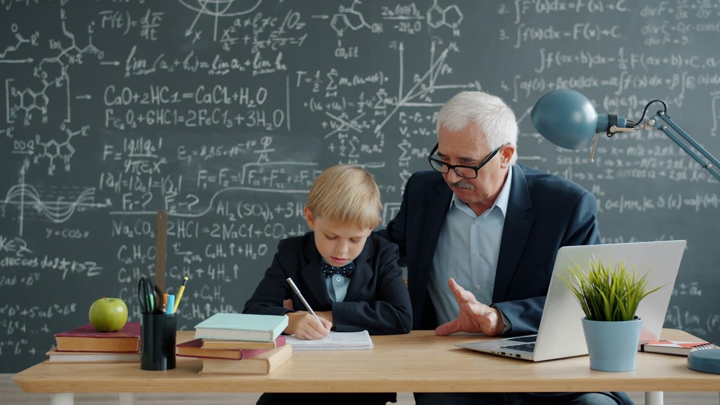 Older man tutoring a young boy with a math worksheet at a desk