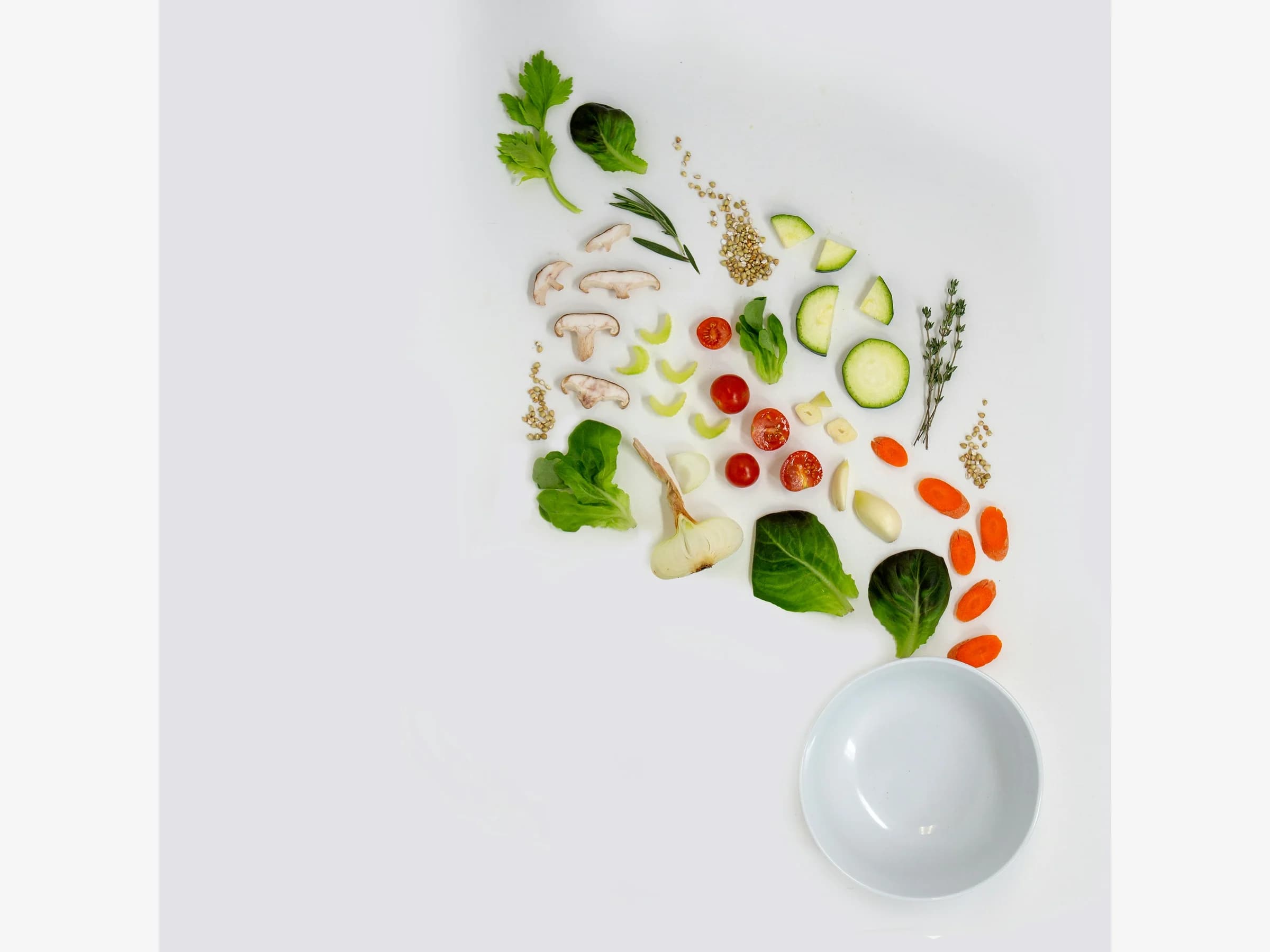 Fresh vegetables arranged beside an empty white bowl on a white table.