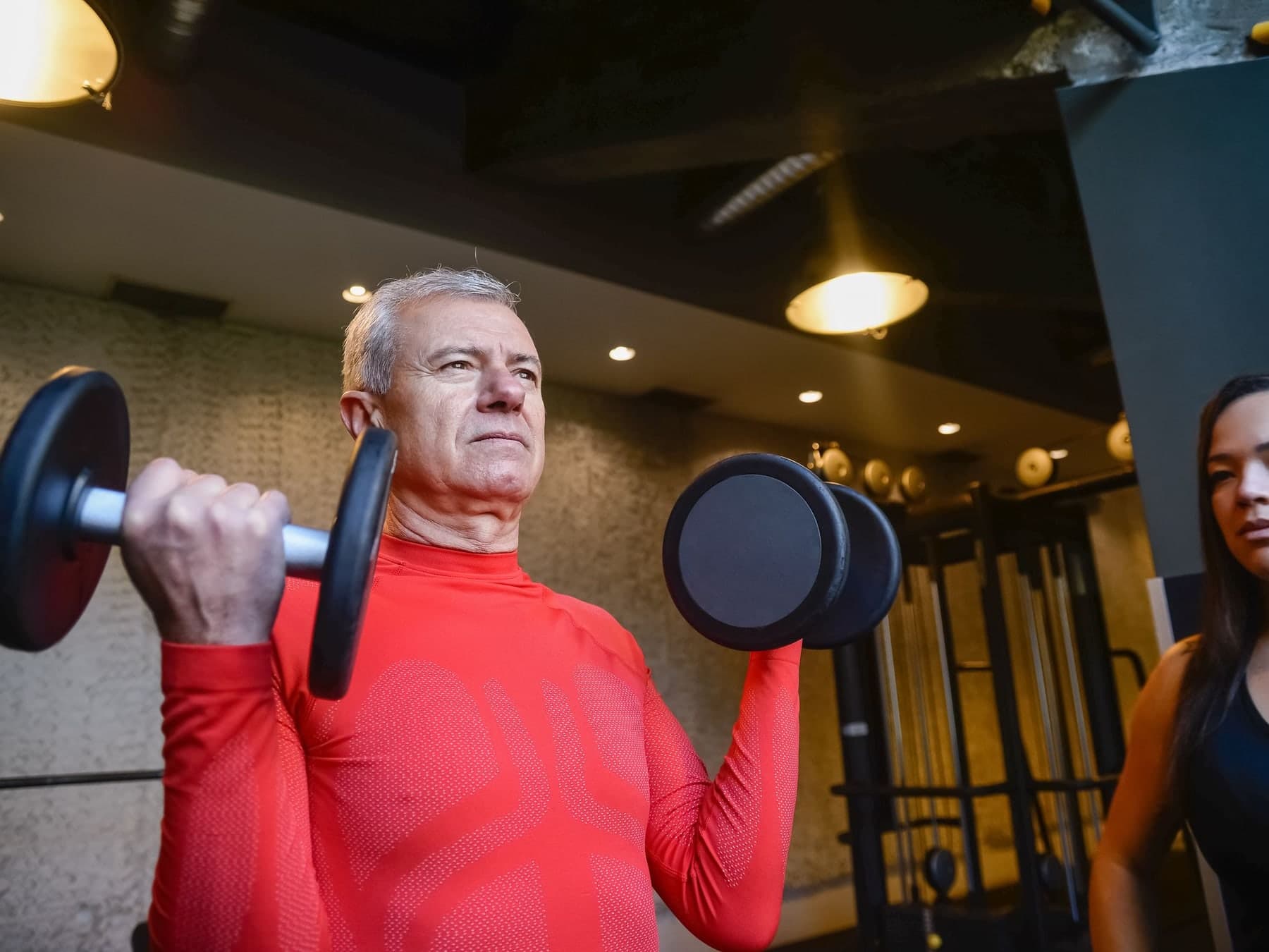 Older man in a red shirt curling dumbbells in a gym while a trainer watches