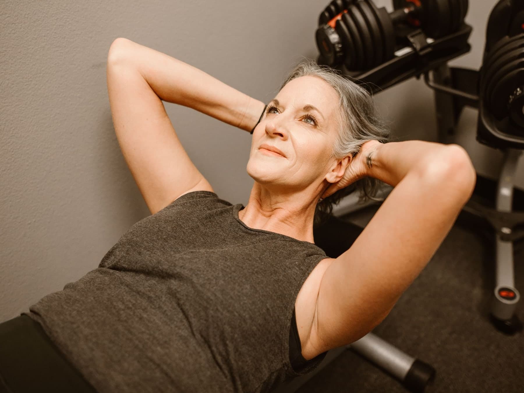 Older woman with gray hair lying on a weight bench with her hands behind her head, an adjustable dumbbell rack behind her