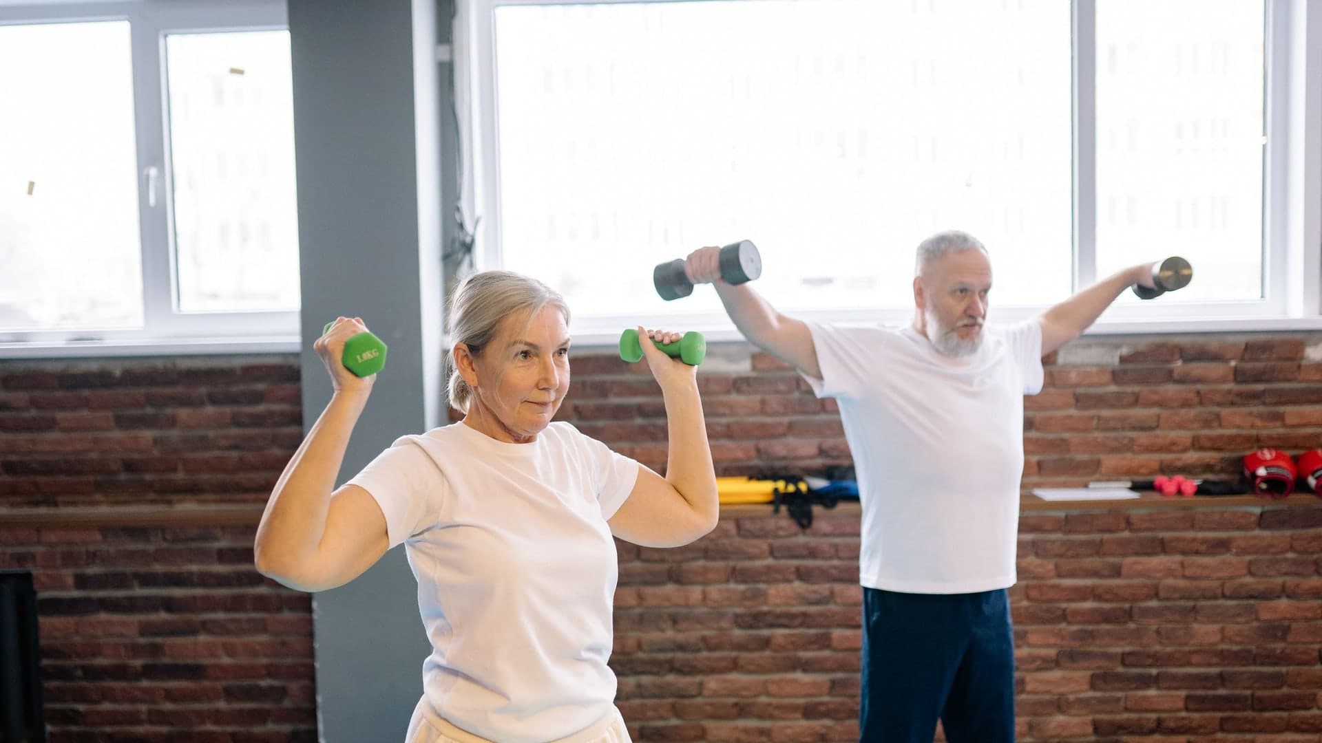 An older man and woman lifting dumbbells side by side in a bright gym